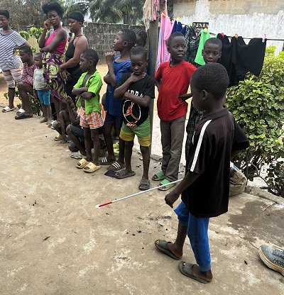 Picture of a little boy practicing walking along the path with his new white cane with kids and adults watching in admiration.