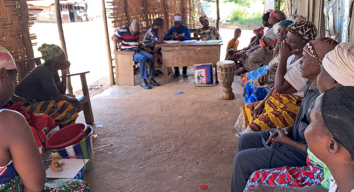 Picture of the community living space for the blind village from a different angle with people sitting in the chairs along the walls.  This picture shows a desk being used by a couple of people.  There are also handmade drums being played by some of the people of the community. 