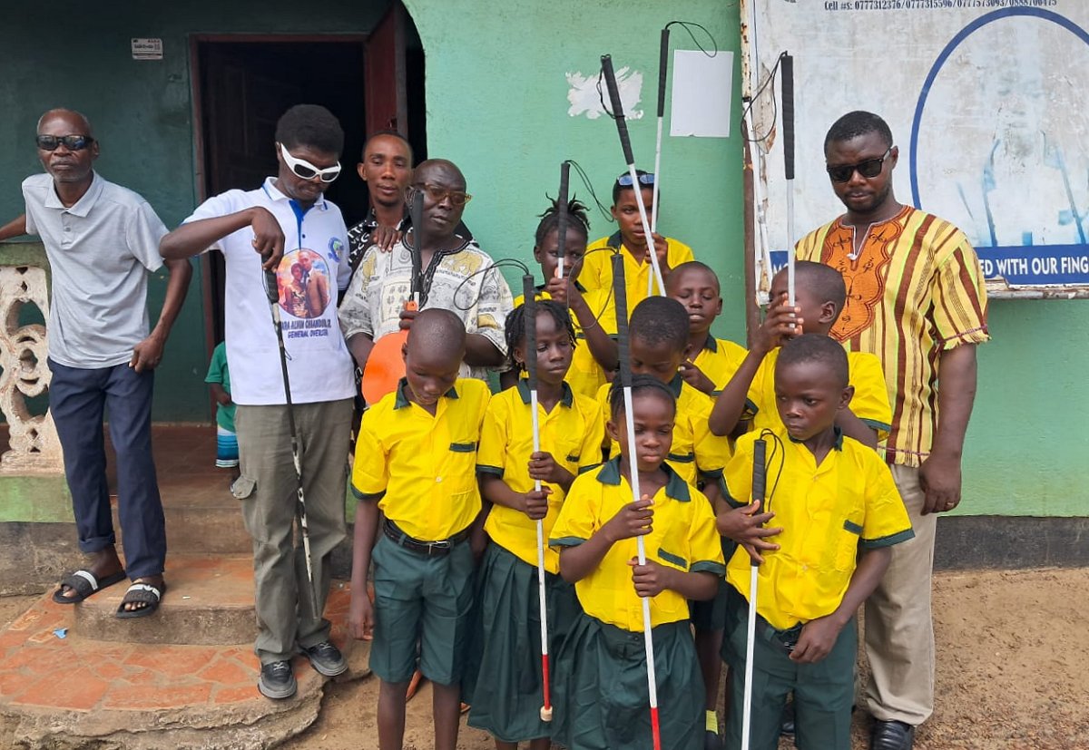 Picture of 9 Students and 5 teachers or care takers standing in front of the school for the blind in Liberia, Africa. They are displaying their white canes.  The students are wearing their school uniforms consisting of yellow and green collared t-shirts and green shorts for boys and skirts for girls.