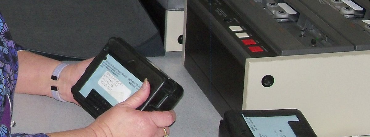 Picture of a Volunteer holding a Cassette Tape mailer and in front of her on the table is a cassette tape duplicator.