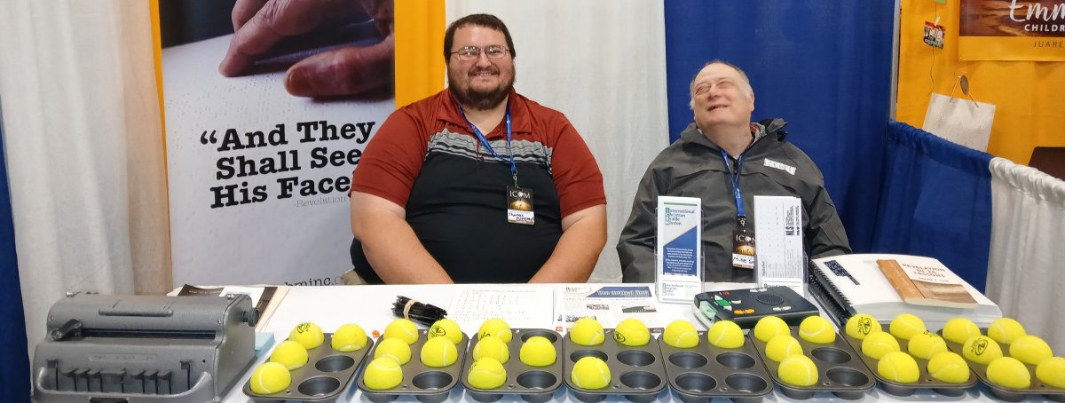Picture of Current President of ICBM Thomas Bleemel and Founder of ICBM Michael Smith.  They are seated behind a display table at the International conference on Missions in Lexington, KY in November 2024.  On the Table are Muffin Tins and tennis balls which are used as a braille learning tool, along with a National Library Services audio player, a perkins brailler, some braille books, a print out of the braille alphabet, the dotted line newsletter, ICBM brochures and business cards.  The braille Learning tool is a muffin tin with 6 spaces and placed in these spaces to represent dots are tennis balls.  These tennis balls can be moved around the cells to practice braille letters and words.  This is an interactive tool for sighted people to be able to spell their name or other words. These tools and using the Perkins Brailler for people to write their name in braille and be able to take it home draw people's attention to the table which gives us the opportunity to share about the Outreach that ICBM does.