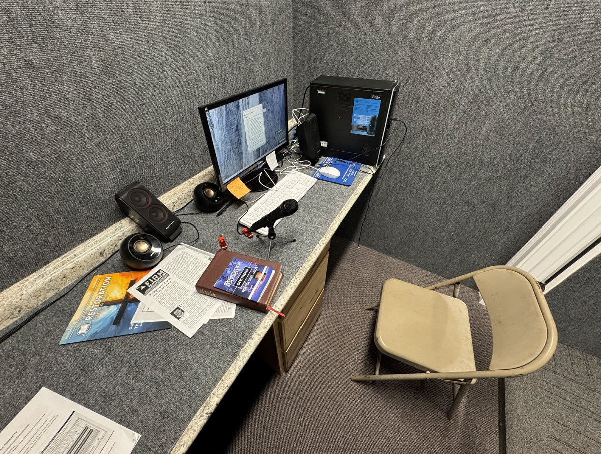 A complete photo of our recording booth at the ICBM offices to record audio for the audio ministry.  On the table is the microphone, a bible with a devotional booklet, some speakers on each side of the computer monitor, and the computer tower along the wall farthest from the photo.  Carpet is on the walls and doors to assist in dimming excess noise.
