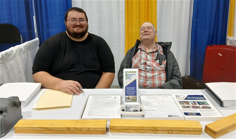 Picture of Current President of ICBM Thomas Bleemel and Founder of ICBM Michael Smith.  They are seated behind a display table at the International conference on Missions in Atlanta, Georgia in November 2025.  On the Table are braille learning tools that Thomas made at home, along with a perkins brailler, some braille books, a print out of the braille alphabet, the dotted line newsletter, ICBM brochures and business cards.  The braille Learning tool is a length of wood with 10 braille cells drilled into it and placed in these cells to represent dots are steel ball bearings.  These ball bearings can be moved around the cells to practice braille letters and words.  This is an interactive tool for sighted people to be able to spell their name or other words. These tools and using the perkins brailler for people to write their name in braille and be able to take it home draw people attention to the table which gives us the opportunity to share about the Outreach that ICBM does.