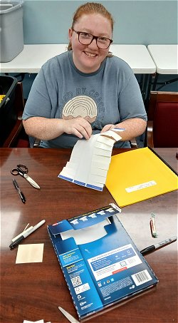 Picture of Wendy who is a volunteer that helps ICBM frequently.  she is sitting at a table, looking up at the camera and smiling while labeling some braille books from the Sharing library at ICBM’s office.
